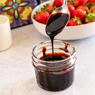 A silver spoon drizzling thick, glossy balsamic glaze into a small glass jar, with a bowl of fresh strawberries in the background.