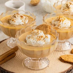 Close up of pumpkin panna cotta in glass cups, each topped with whipped cream and cookie crumbs, served on a wood slab with cinnamon sticks and gingersnap cookies.