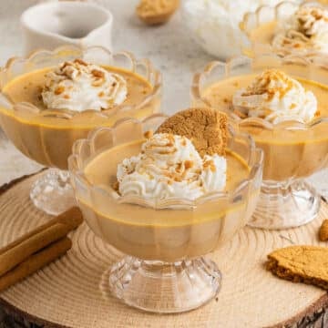 Close up of pumpkin panna cotta in glass cups, each topped with whipped cream and cookie crumbs, served on a wood slab with cinnamon sticks and gingersnap cookies.
