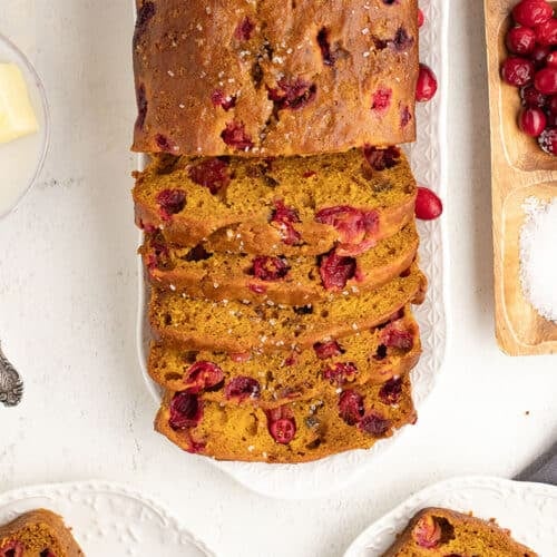 Close up overhead shot of sliced pumpkin cranberry bread on a platter, surrounded by cranberries and butter.