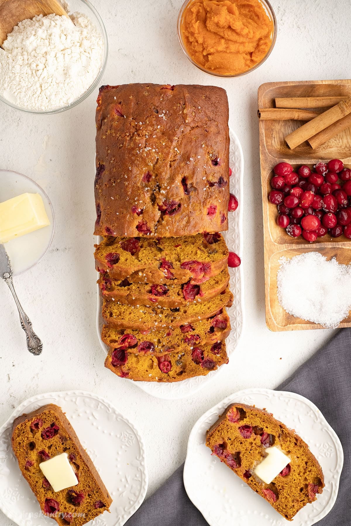 Overhead shot of sliced pumpkin cranberry bread on a platter, surrounded by ingredients and two buttered slices on plates.
