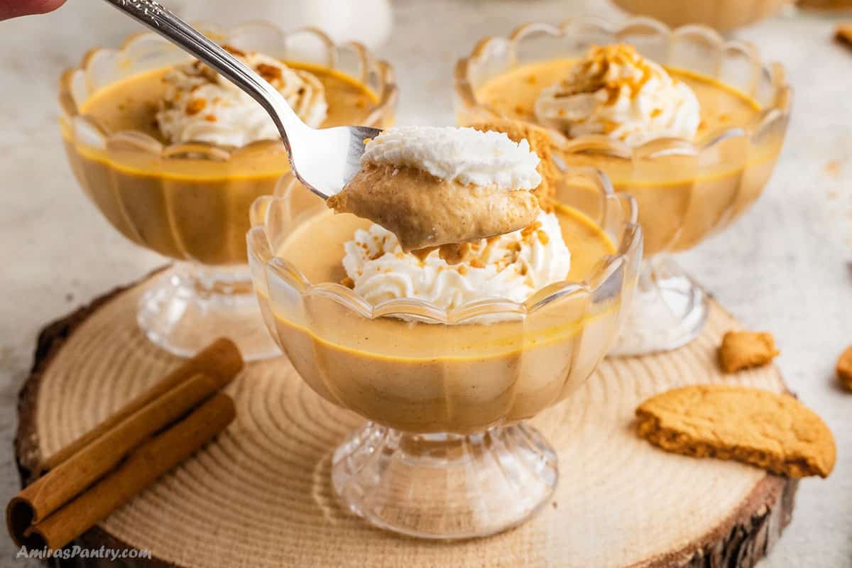 A spoon lifting a creamy bite of pumpkin panna cotta topped with whipped cream from a glass dish, with more servings and cinnamon sticks in the background.