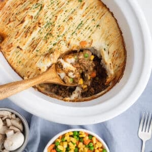 A wooden spoon scooping a serving of cottage pie out of a white slow cooker.