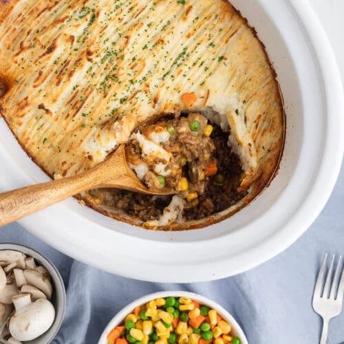A wooden spoon scooping a serving of cottage pie out of a white slow cooker.
