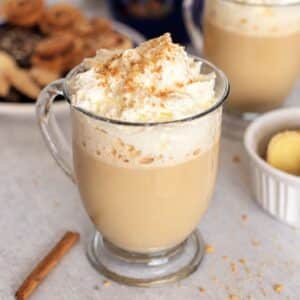 Glass mug of ginger latte topped with whipped cream and crumb sprinkles, with a cinnamon stick beside it.