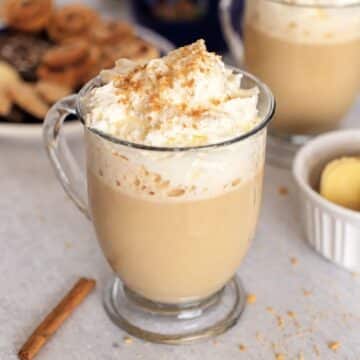 Glass mug of ginger latte topped with whipped cream and crumb sprinkles, with a cinnamon stick beside it.