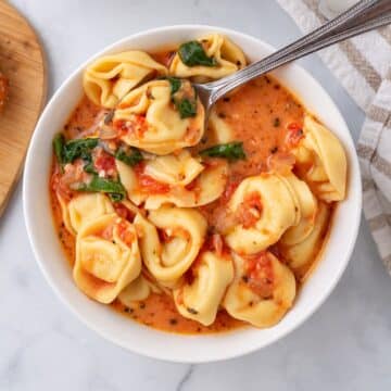 Bowl of creamy tomato tortellini soup with spinach and a spoon scooping up a tortellini.