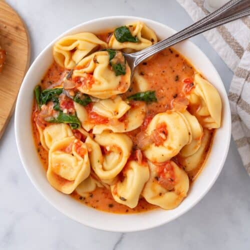Bowl of creamy tomato tortellini soup with spinach and a spoon scooping up a tortellini.