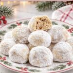Stack of powdered sugar pecan snowball cookies on a holiday plate, one with a bite taken out, surrounded by pine branches and red berries.