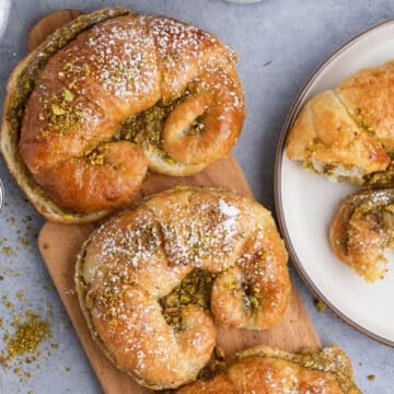 Pistachio croissants dusted with powdered sugar and crushed pistachios on a wooden board, with a torn piece on a plate nearby.