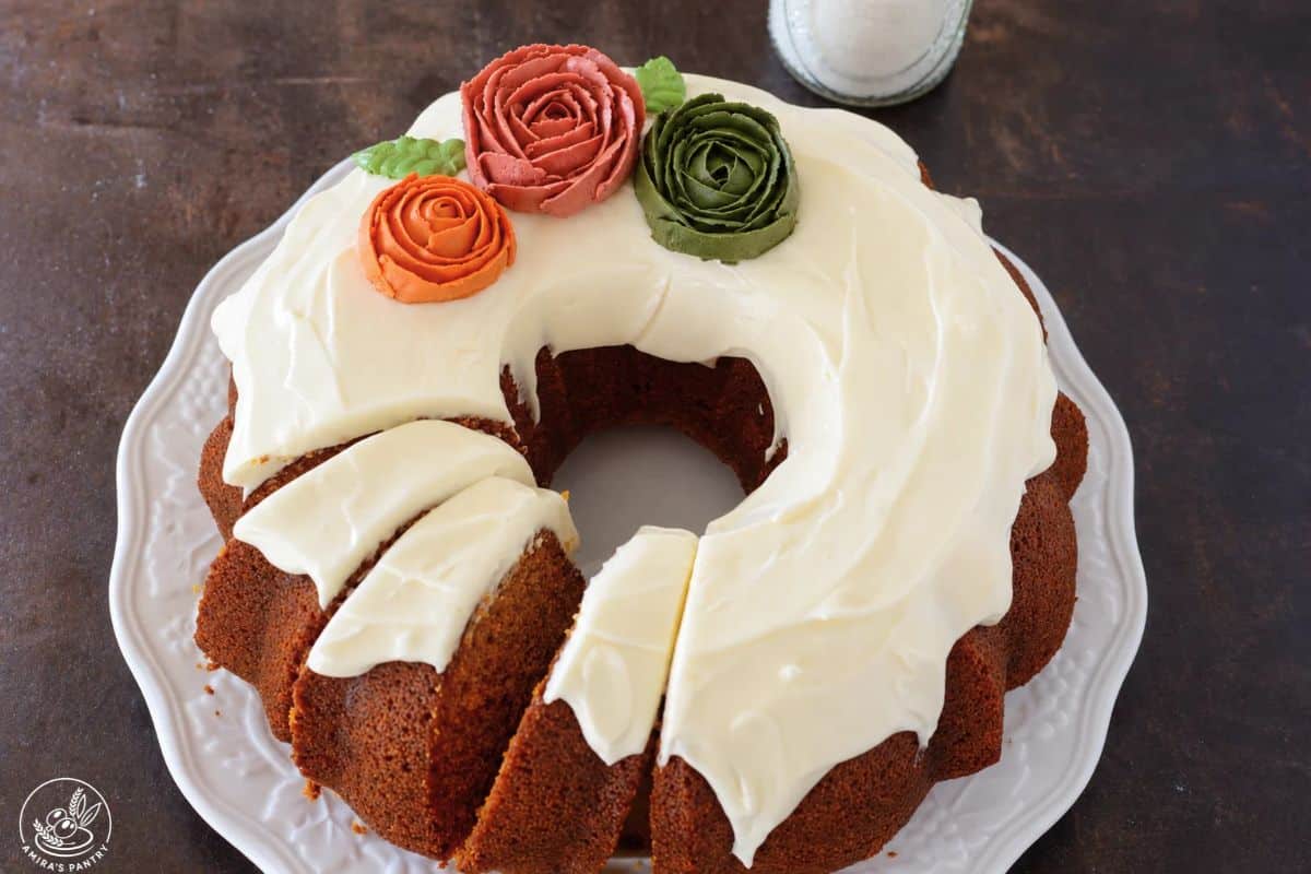 Frosted pumpkin bundt cake with orange, coral, and green frosting roses, partly sliced on a white plate.